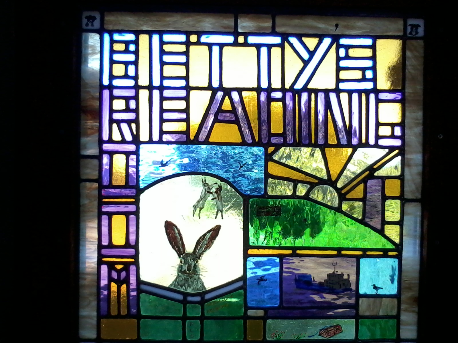 A stained glass window, which includes the words 'Betty's Reading Room' in purple on a yellow background. There is a painting of a rabbit in one element of the glass, while another has a sunrise, and others have nautical elements including a ferry, seabirds, and a lobster pot.