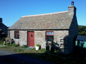A stone-built, slate-roofed cottage under a blue sky. It is only one-storey tall. There is a single red door, and one window to each side of the door, with red wooden frames. There is grass in front of the cottage, and a few plants in pots.