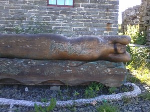 A wooden sculpture of a lady lying on her side. It is situated outside, on grass, by a stone wall.